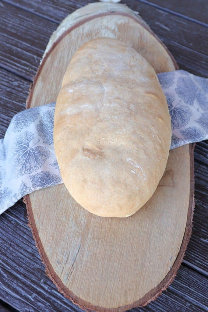An oblong loaf of white hearth bread sits on a napkin that is draped over a wooden board as seen from above.
