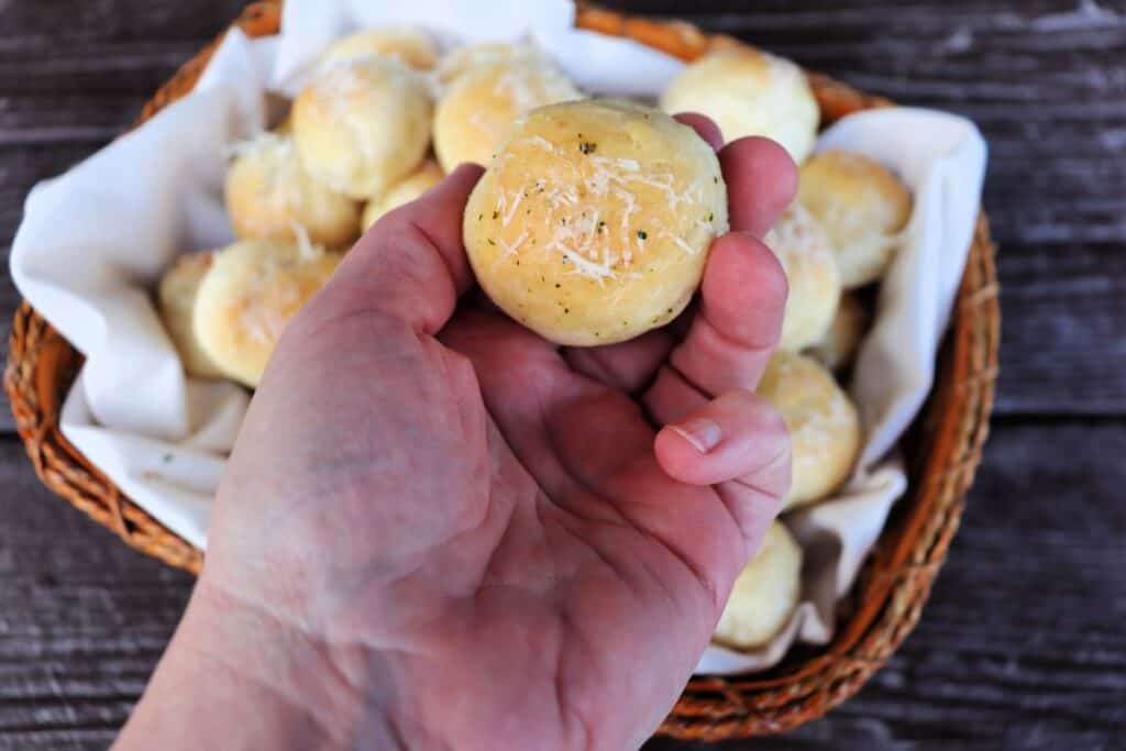 A hand holding a bread ball covered in grated parmesan cheese with a basket of more behind it.