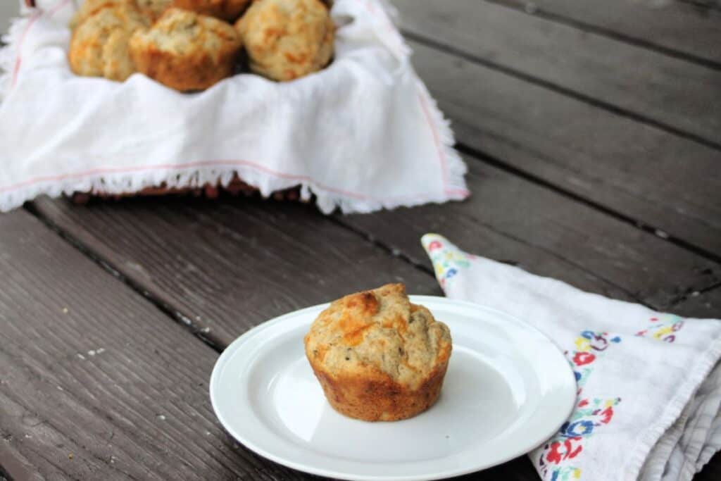 A cheddar rye muffin on a plate with a napkin to the right. In the background is a linen lined basket full of more muffins.