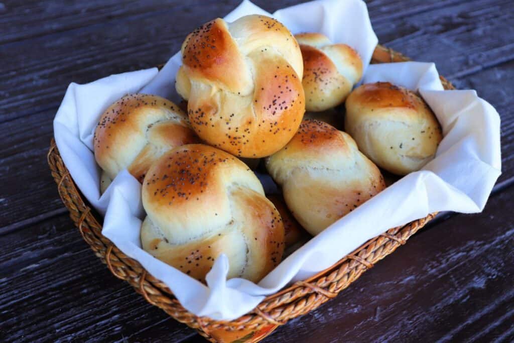 A linen lined basket of knotted challah rolls sits on a table.