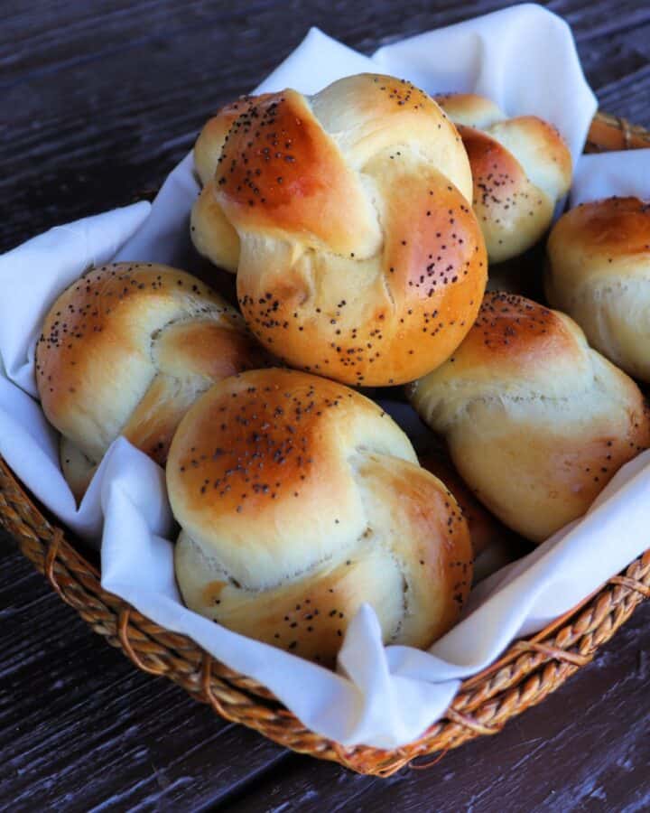 A linen lined basket full of poppy seed topped knotted challah rolls.