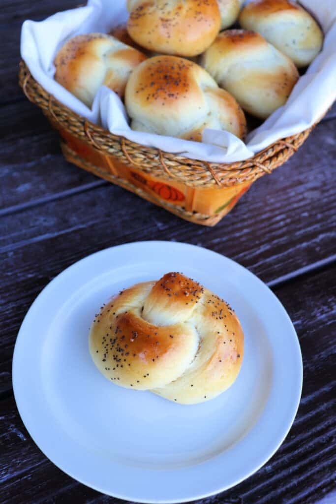 A poppy seed knotted challah roll sits on a plate with a linen lined basket full of more rolls in the background.