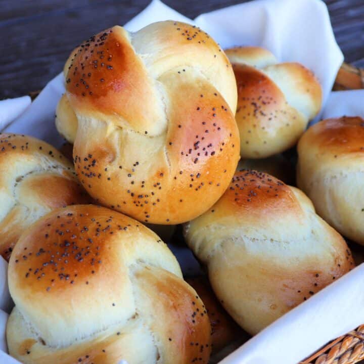 A poppy seed covered challah roll sitting on top of more rolls in a linen lined basket.