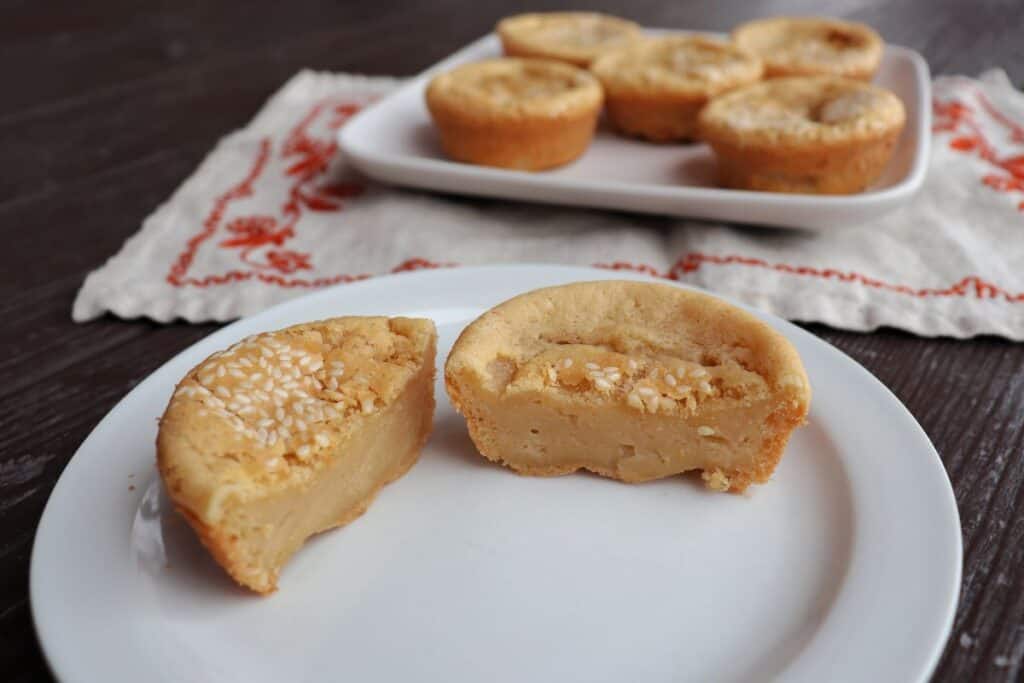 A sesame topped mochi muffin sliced in half sits on a plate. More muffins sit on a platter sitting on a table runner in the background.