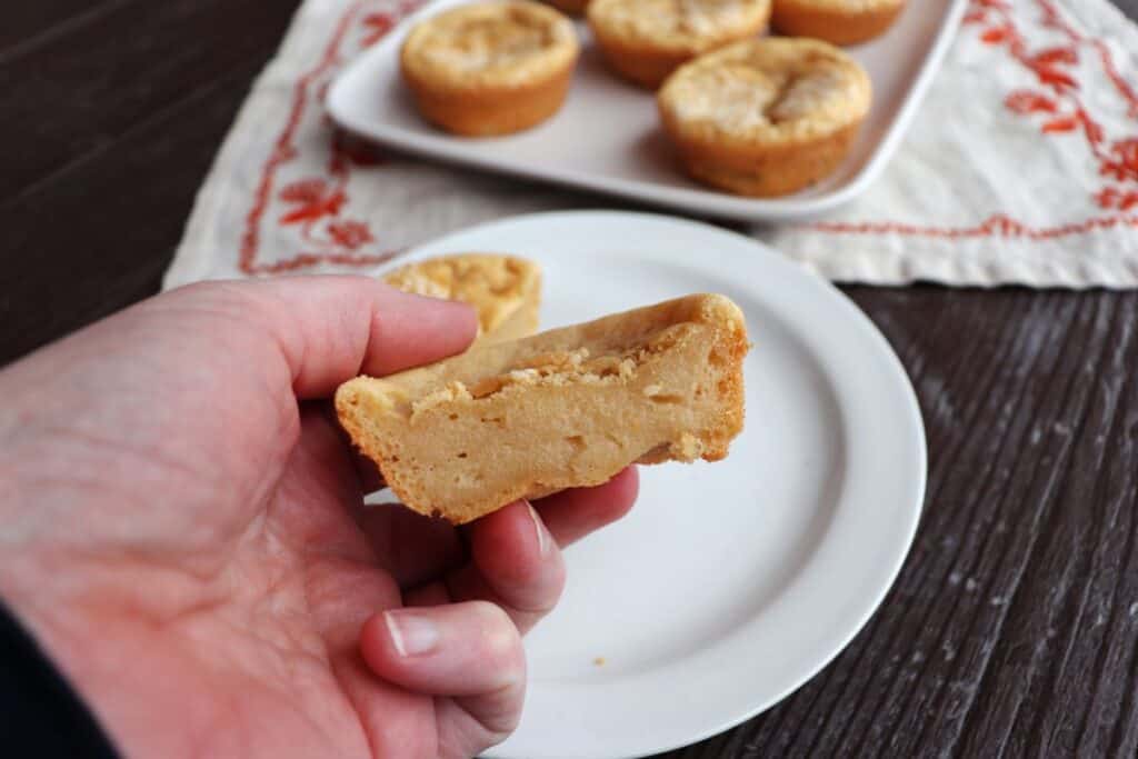 A woman's hand holding half of a mochi muffin exposing the inside. A plate and more muffins in the background