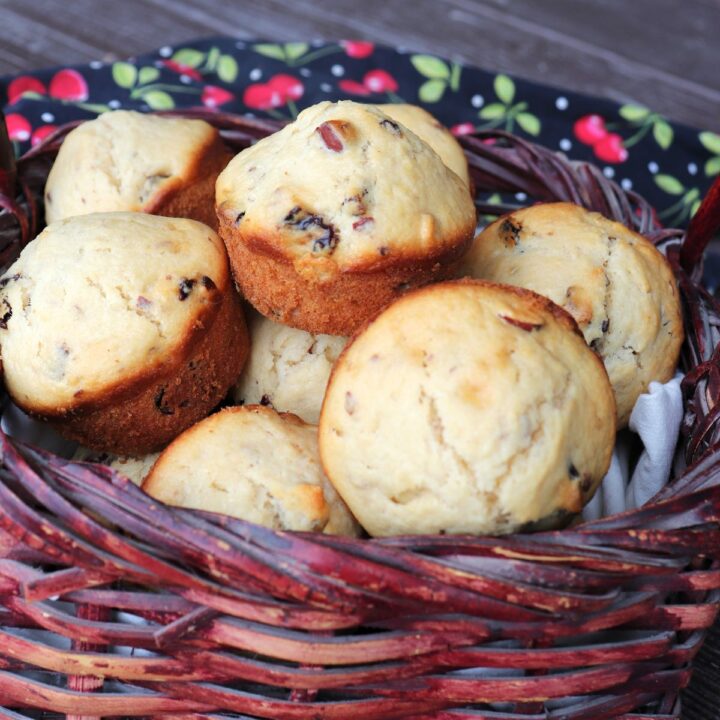 A basket full of dried cherry muffins sits on a table with a cherry printed cloth in the background.