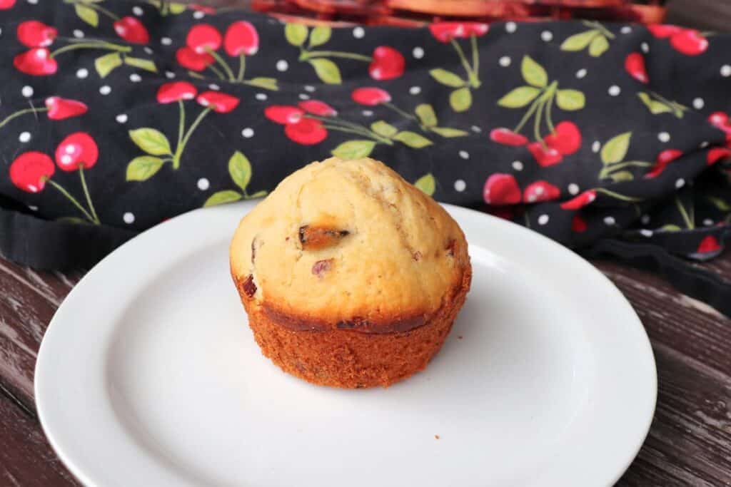 A dried cherry muffin sits on a plate with a cherry printed cloth in the background.