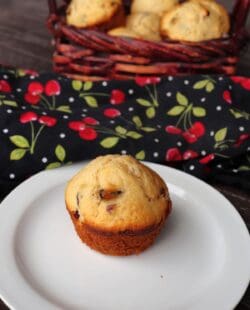 A dried cherry muffin sits on a plate with a basket of more muffins and a cherry printed cloth in the background.