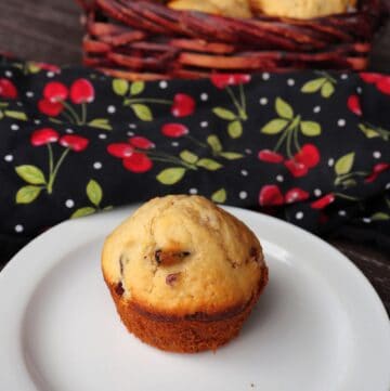 A dried cherry muffin sits on a plate with a basket of more muffins and a cherry printed cloth in the background.