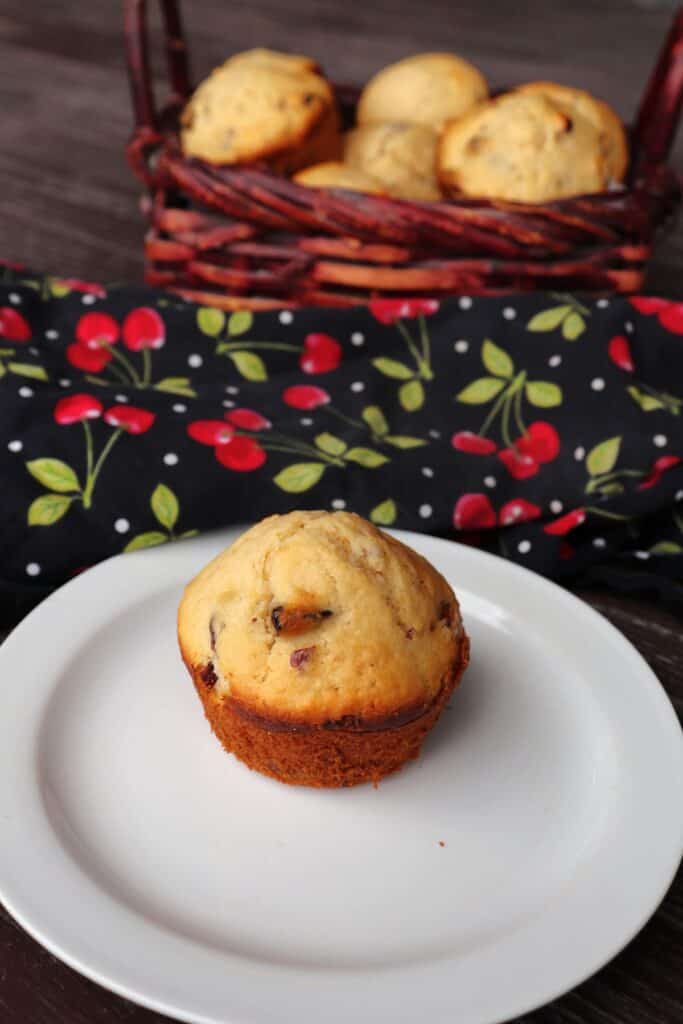 A dried cherry muffin sits on a  plate with a basket of more muffins and a cherry printed cloth in the background.