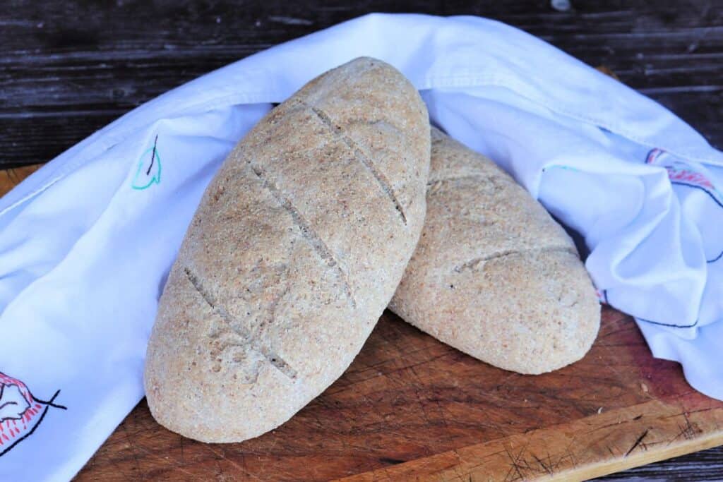 A loaf of whole wheat Italian bread sits on top of another, both surrounded by a white cloth on a wood board.