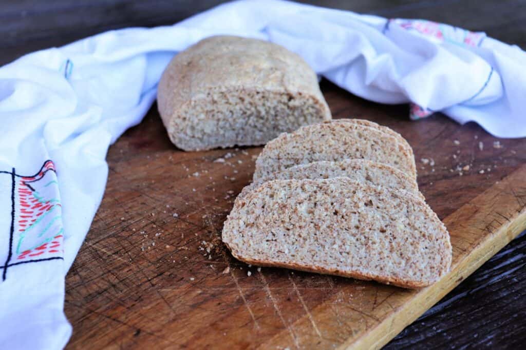 Slices of whole wheat Italian Bread sit on a board with remaining loaf and white cloth in the background.