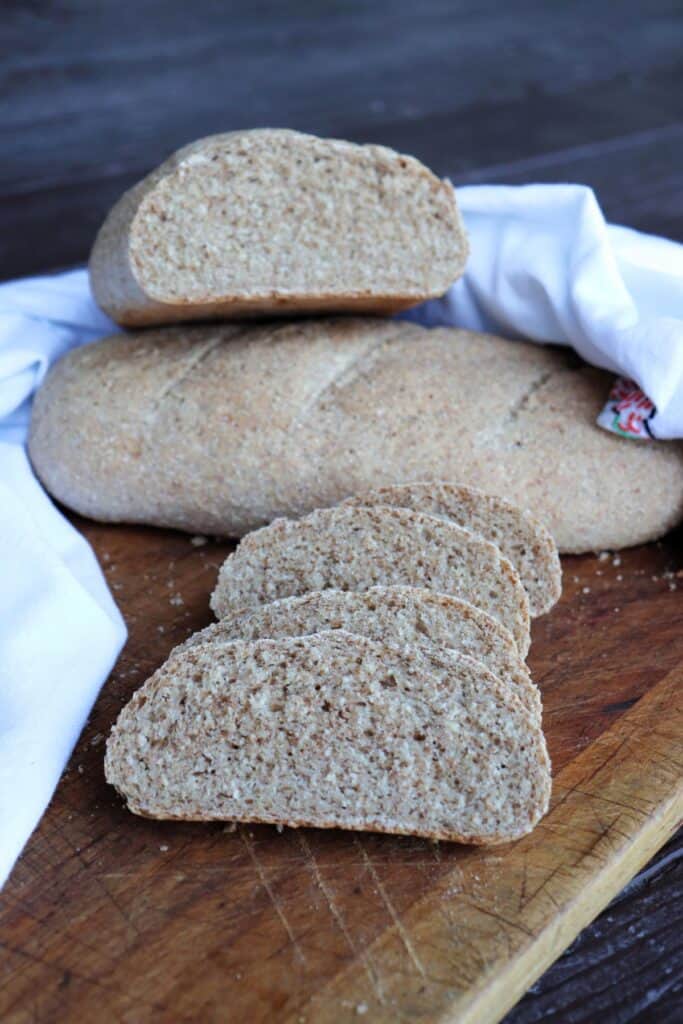 Slices of whole wheat Italian Bread sit on a board with the remaining loaf of bread sitting on another loaf behind them. All is surrounded by a white cloth.