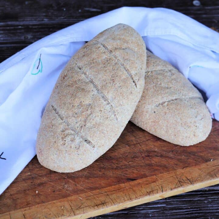 A loaf of whole wheat Italian bread sits on top of another on a board, a white cloth surrounding both.