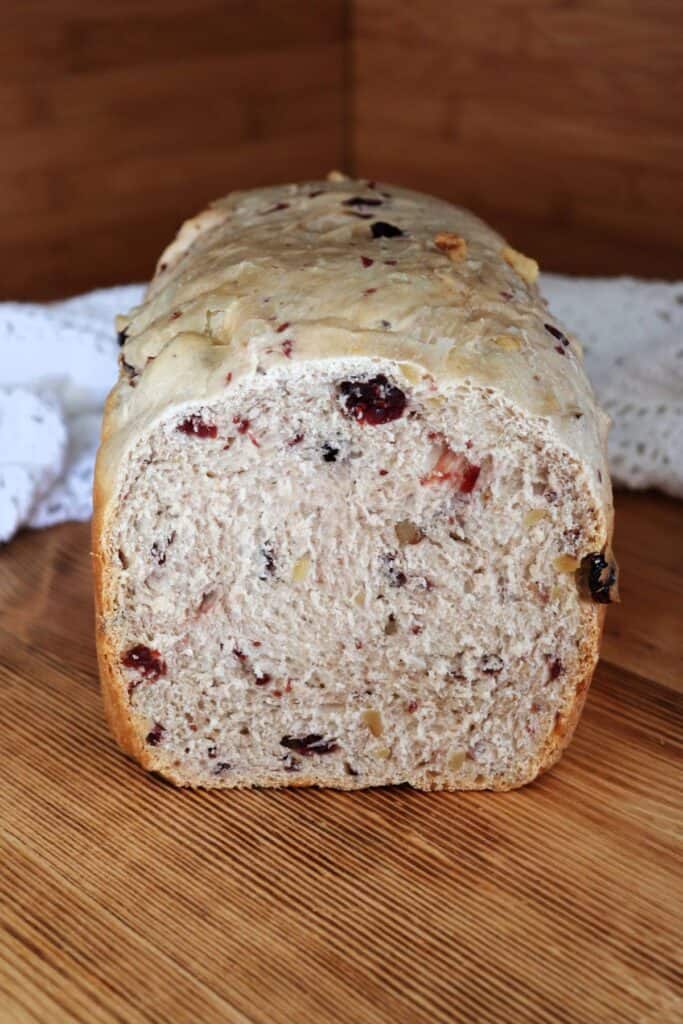 A loaf of bread with tthe end cut off exposing the inside studded with dried cranberries and walnuts sits on a board with a white cloth behind it.