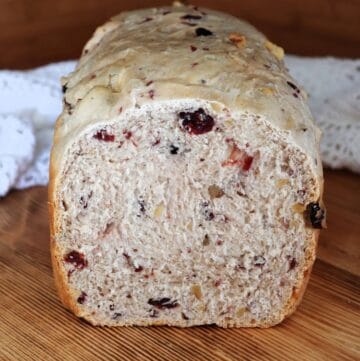 A loaf of bread with tthe end cut off exposing the inside studded with dried cranberries and walnuts sits on a board with a white cloth behind it.