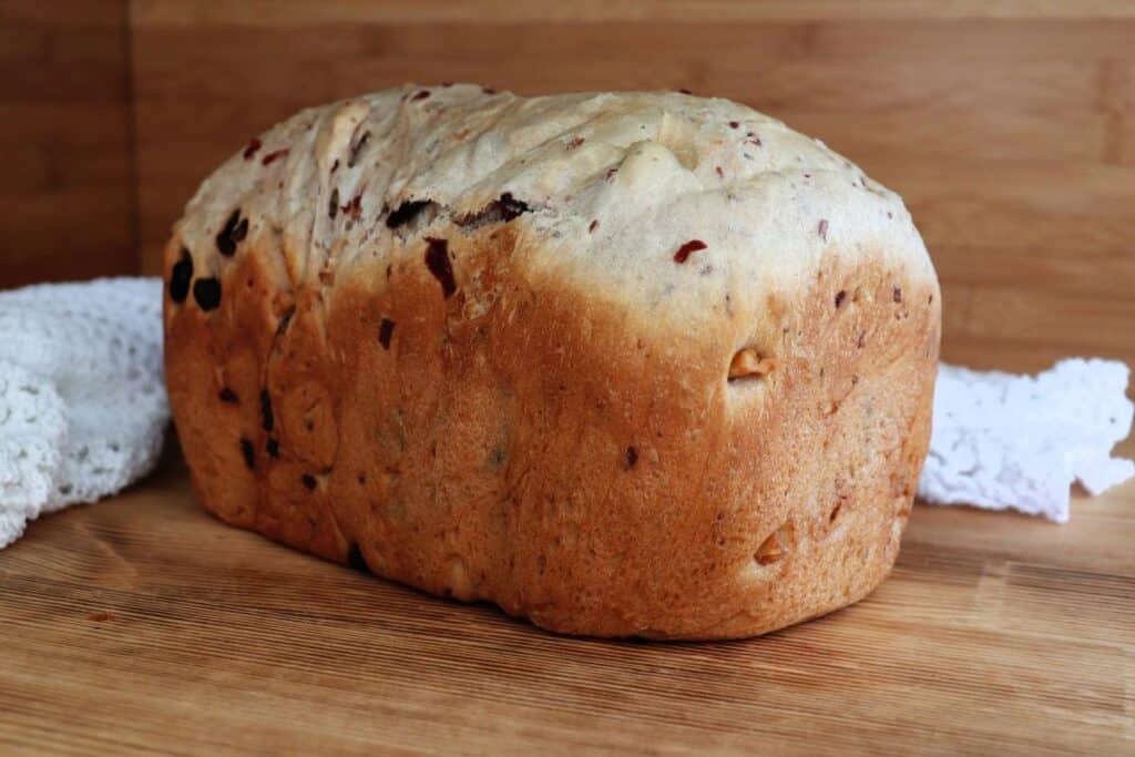 A loaf of bread studded with walnuts and dried cranberries sits on a board with a white cloth behind it. 