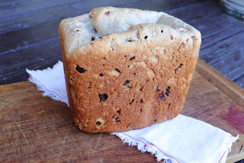 A loaf of cranberry walnut bread sits on top of a cloth napkin draped across a wood cutting board.