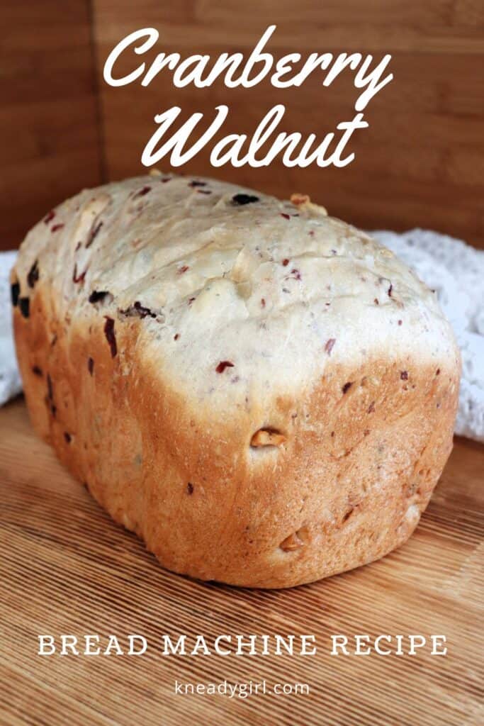 A loaf of bread studded with walnuts and dried cranberries sits on a board with a white cloth behind it. Text overlay reads: Cranberry Walnut Bread Machine Recipe. 