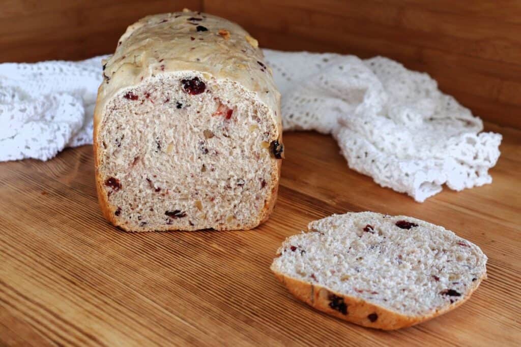 A loaf of bread with tthe end cut off exposing the inside studded with dried cranberries and walnuts sits on a board with a white cloth behind it.
