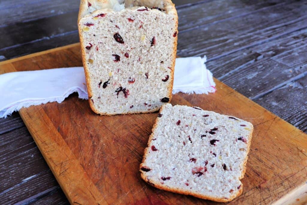 A slice of cranberry walnut bread sits on a board with the remaining loaf sitting on a cloth napkin behind it. 