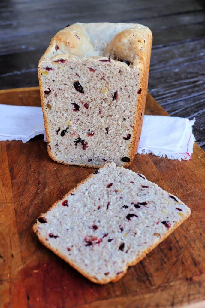 A slice of cranberry walnut bread sits on a board with the remaining loaf in the background sitting on a cloth napkin.