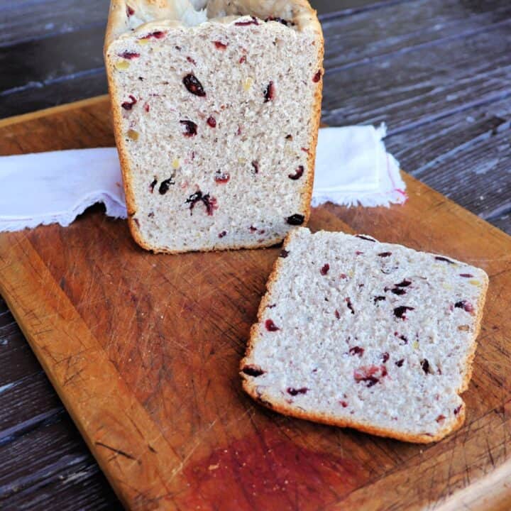 A slice of cranberry walnut bread sits on a board with the remaining loaf in the background sitting on a cloth napkin.