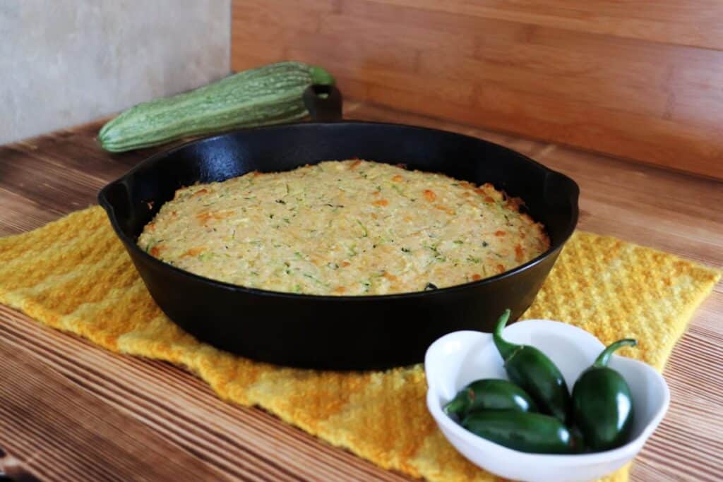 A cast iron skillet full of zucchini cornbread sits on a yellow runner. A bowl of hot peppers in the foreground. A zucchini in the background. 