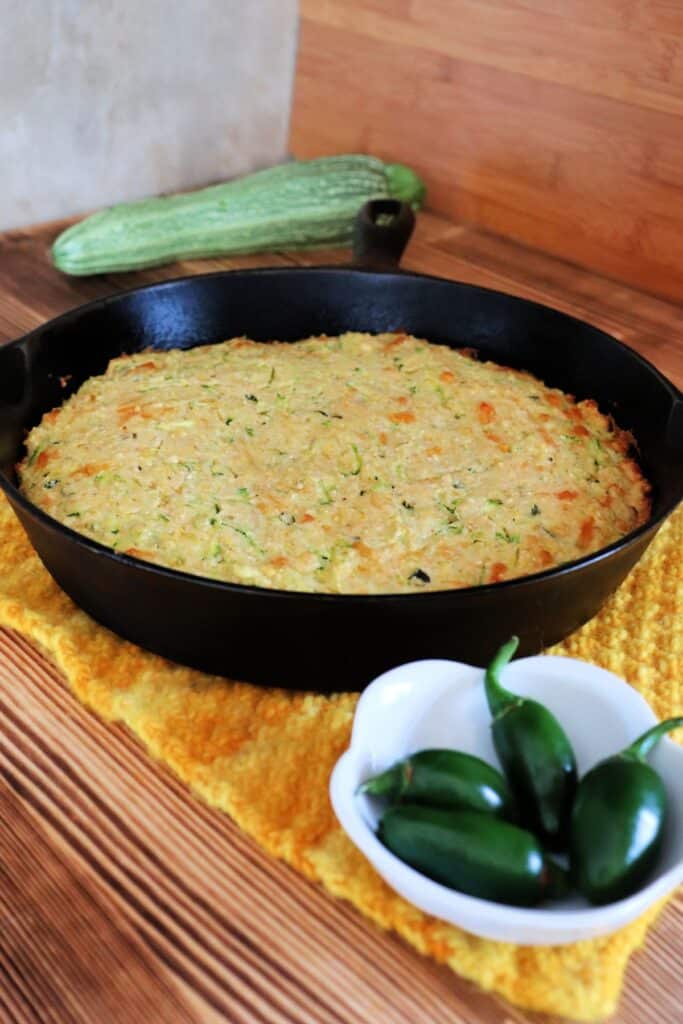 A cast iron skillet full of cornbread sits on a yellow cloth. A bowl of hot peppers sits in front of the skillet, a zucchini sits in the background. 