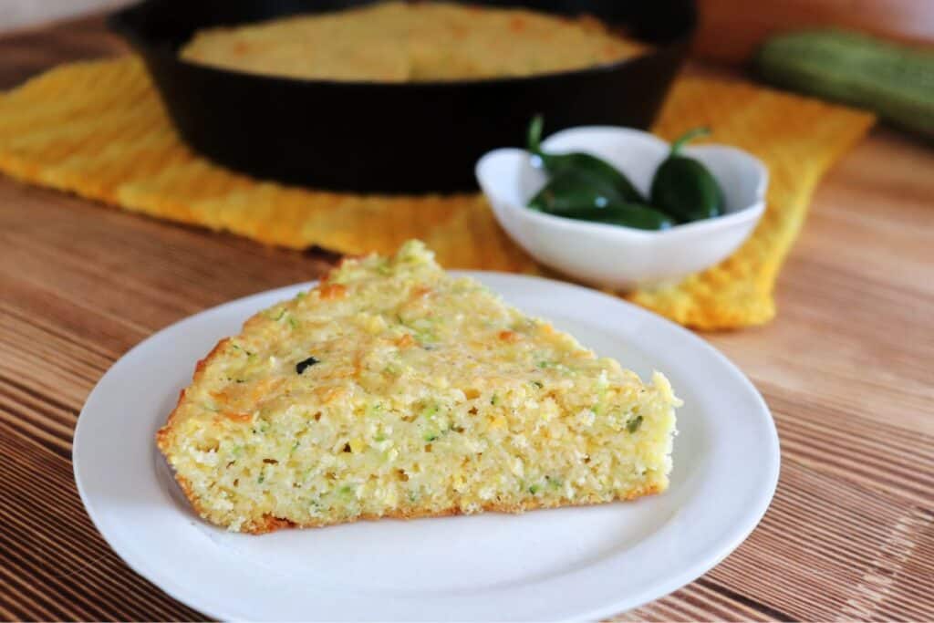 A slice of zucchini cornbread on a plate with a bowl of hot peppers and cast iron skillet with more cornbread in the background.