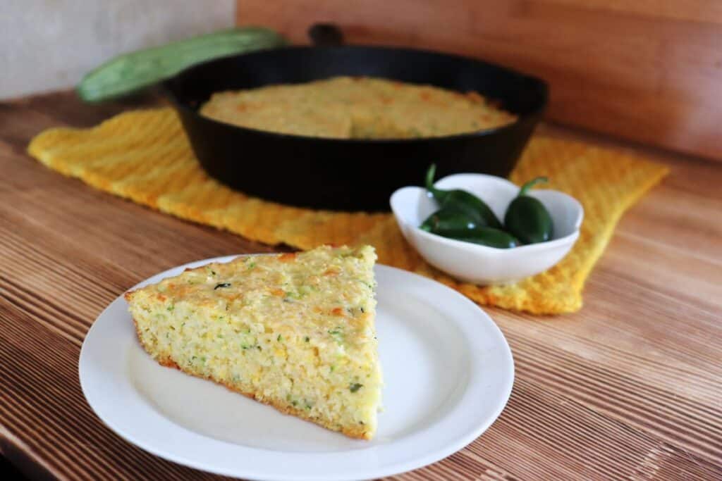 A slice of zucchini cornbread on a plate with a bowl of hot peppers and cast iron skillet with more cornbread in the background.