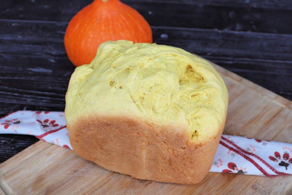 A loaf of bread sits on a cloth draped board with a fresh pumpkin in the background.