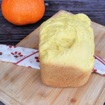 A loaf of bread sits on a cloth draped board with a fresh pumpkin in the background.