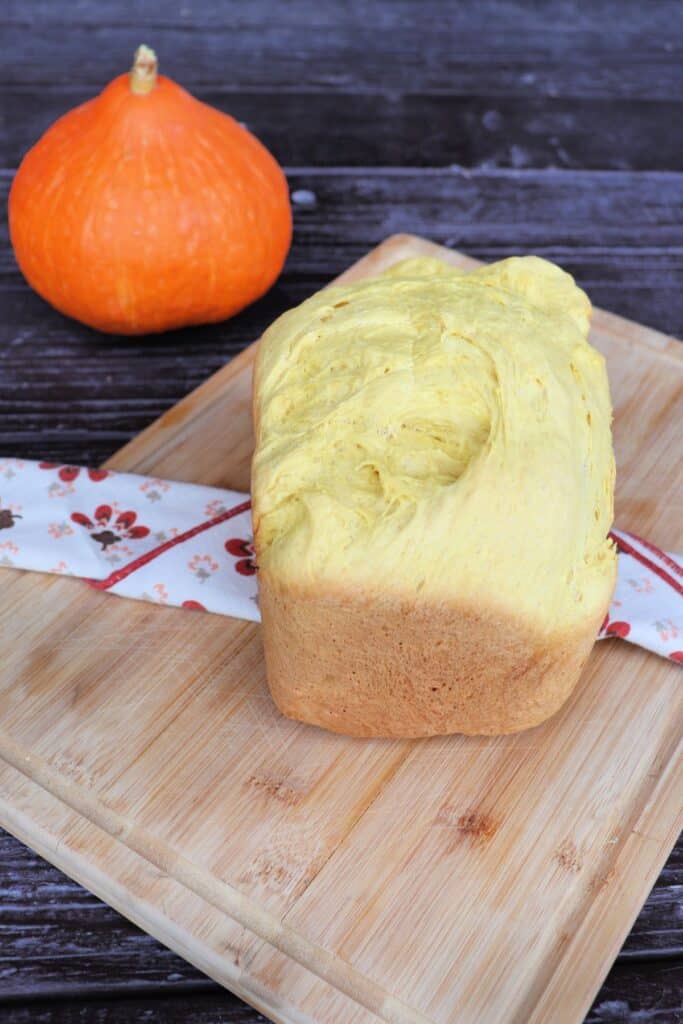 A loaf of bread sits on a cloth draped board with a fresh pumpkin in the background.