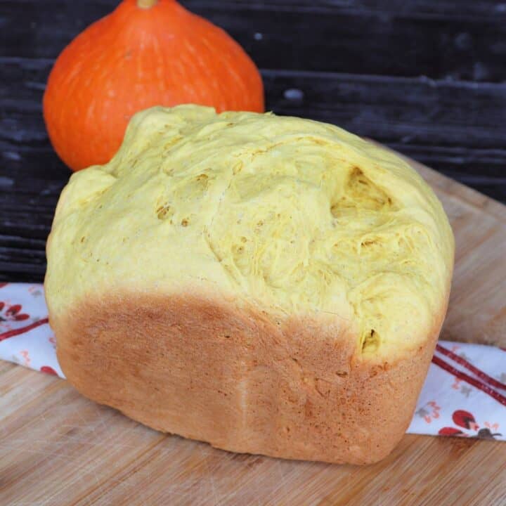 A loaf of pumpkin bread sits on a cloth draped board with a fresh pumpkin in the background.