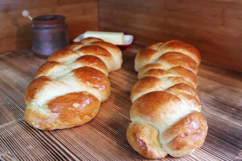 2 loaves of swiss egg bread sit next to each other on a board. A honey pot and butter dish in the background. 