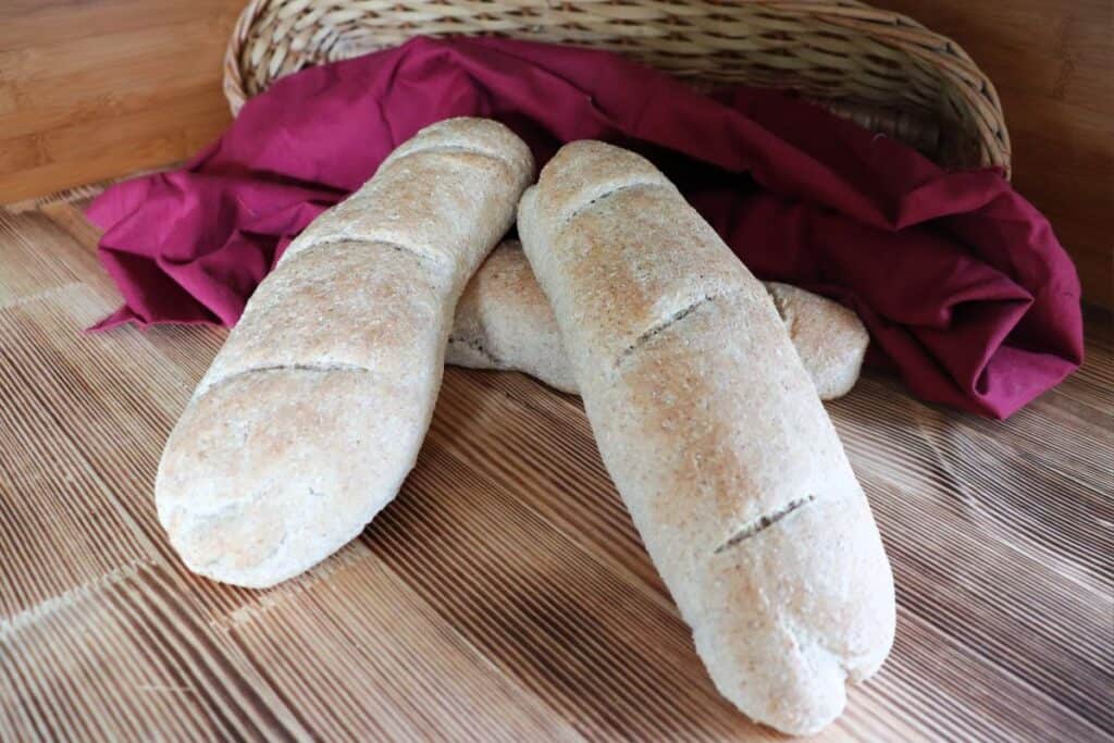 3 whole wheat baguettes sit on a wooden board - a red cloth and basket in the background.