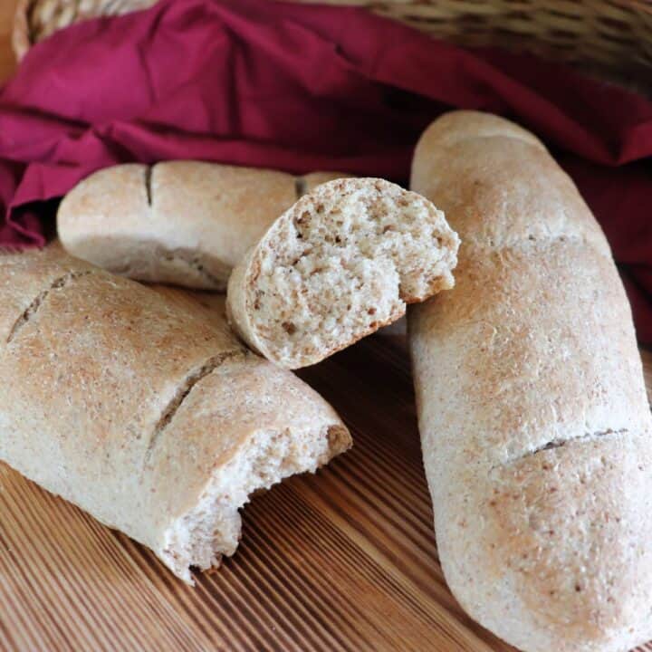 A piece of torn bread sits on top of 3 other whole wheat baguettes on a board with a red cloth in the background.