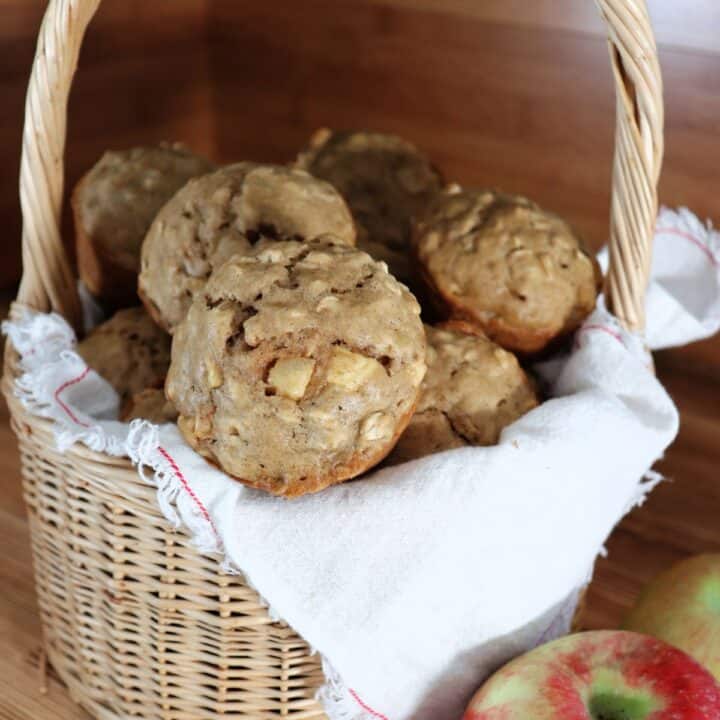 A white linen lined basket full of muffins with fresh apples sitting next to it.