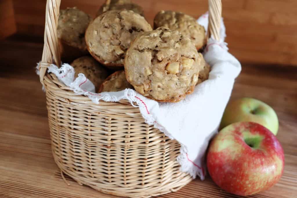 A basket of full of apple oatmeal muffins sits on a table with fresh apples beside it. 