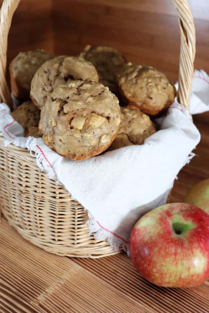 A basket of full of apple oatmeal muffins sits on a table with fresh apples beside it. 