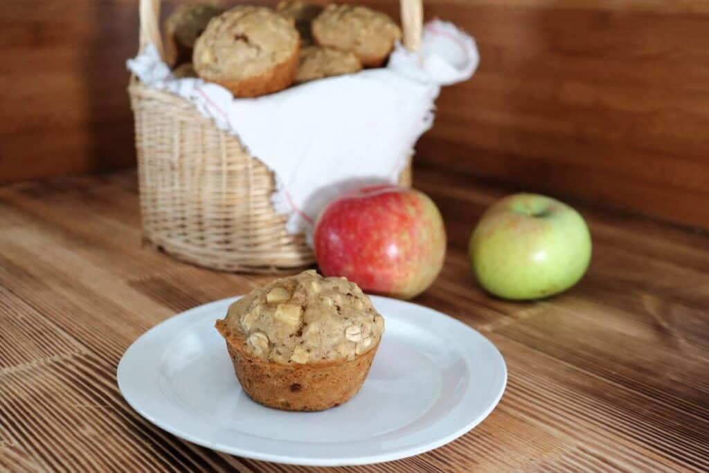 A muffin sits on a plate with white cloth lined basket full of more muffins and fresh apples in the background. 