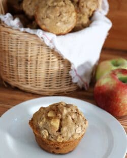 A basket of full of apple oatmeal muffins sits on a table with fresh apples beside it.