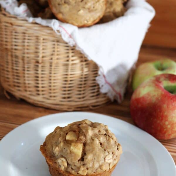A basket of full of apple oatmeal muffins sits on a table with fresh apples beside it.