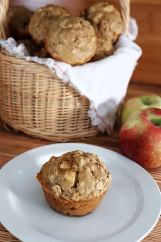 A muffin sits on a plate with white cloth lined basket full of more muffins and fresh apples in the background. 