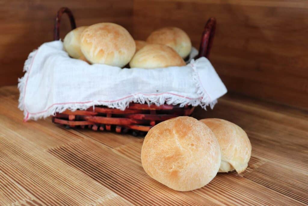 2 dinner rolls sit on a table with a cloth lined basket full of more rolls behind them.