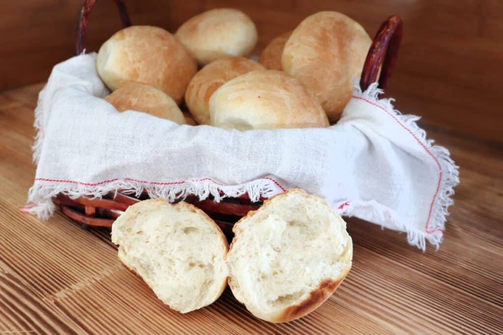 A bread roll is torn in half and sits on a table with insides exposed, a basket of more rolls sits behind it. 