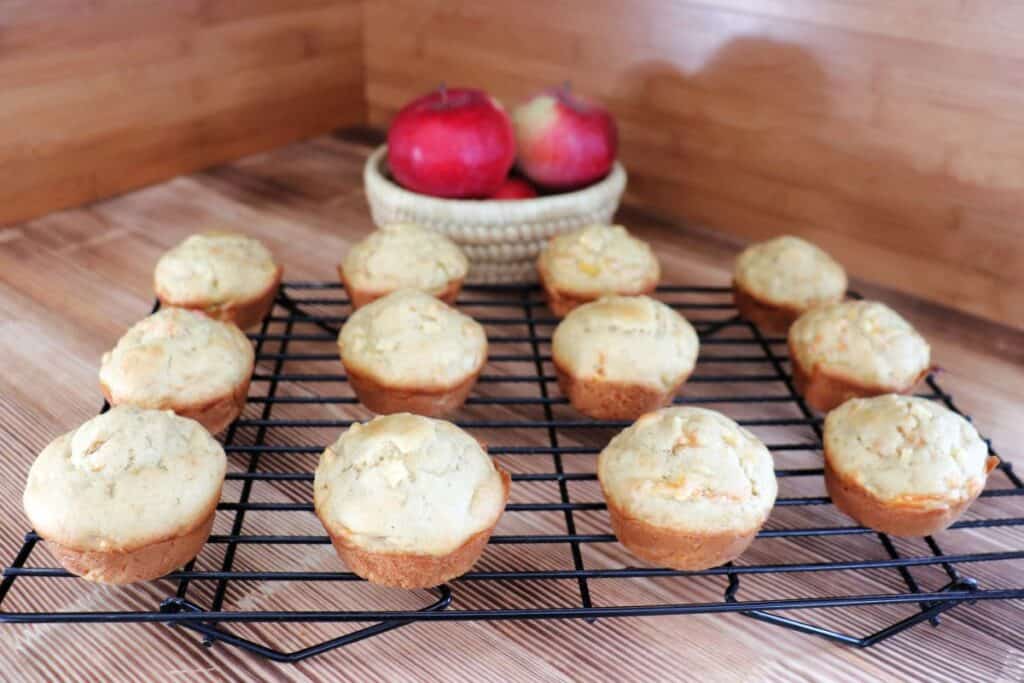 A dozen muffins sit on a wire cooling rack with a basket of fresh apples behind it. 