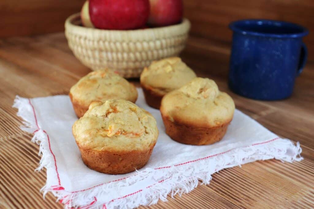 4 carrot apple muffins sitting on a white napkin. In the background is a blue tin coffee cup and a basket of fresh red apples.