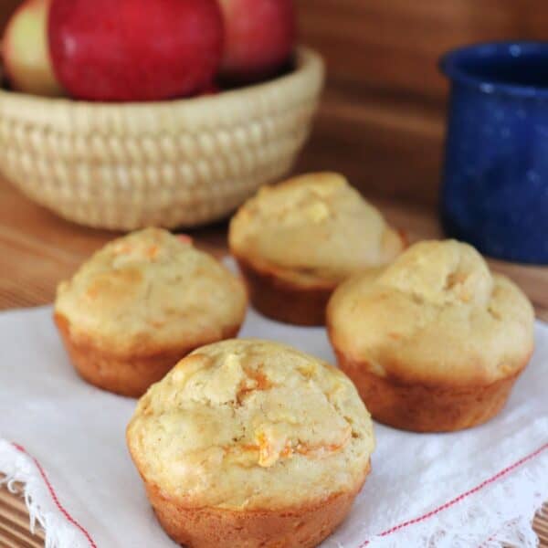 4 carrot apple muffins sitting on a white napkin. In the background is a blue tin coffee cup and a basket of fresh red apples.